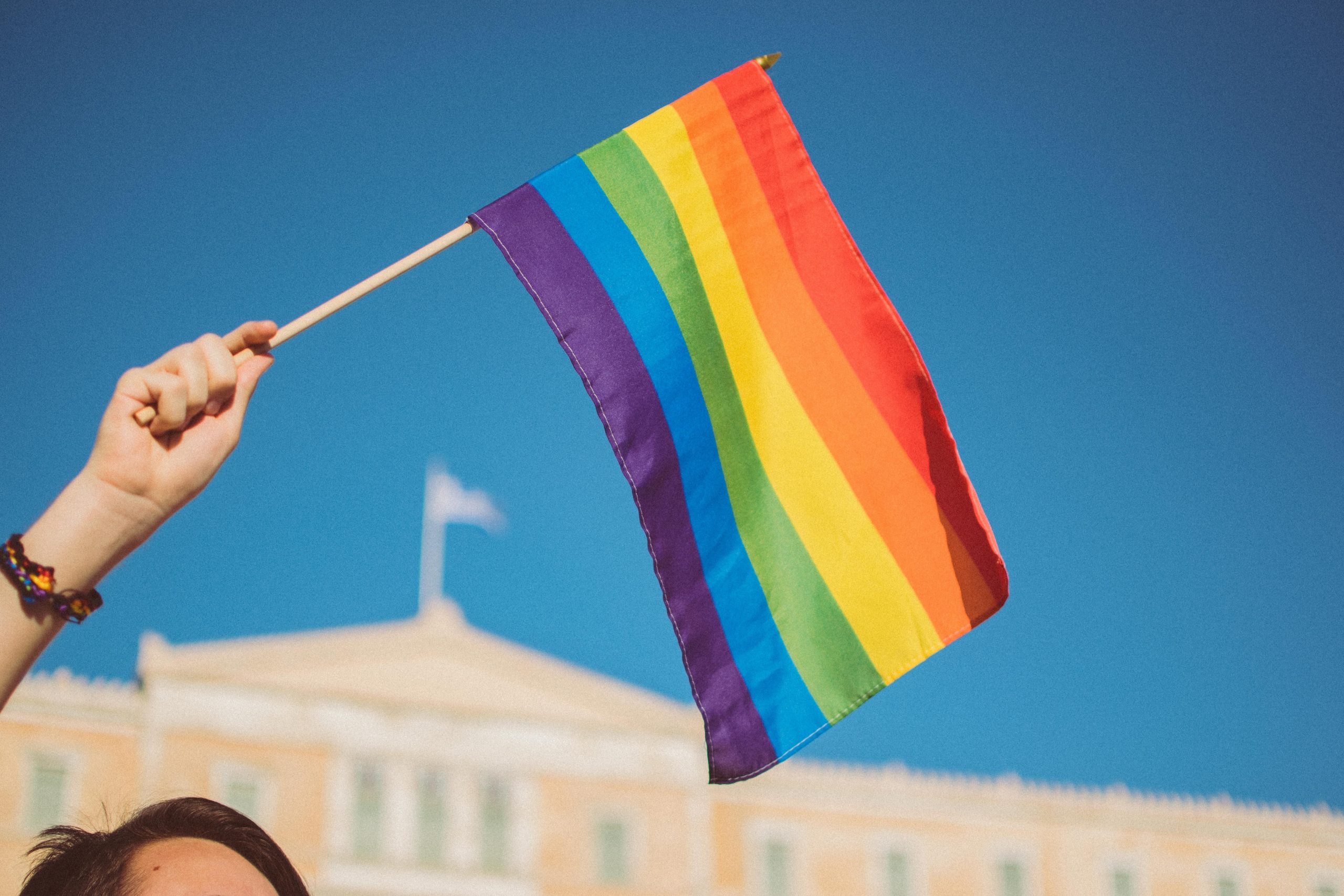 A person's hand holding a LGBTQIA rainbow flag with a blue sky in the background and the top of a house.